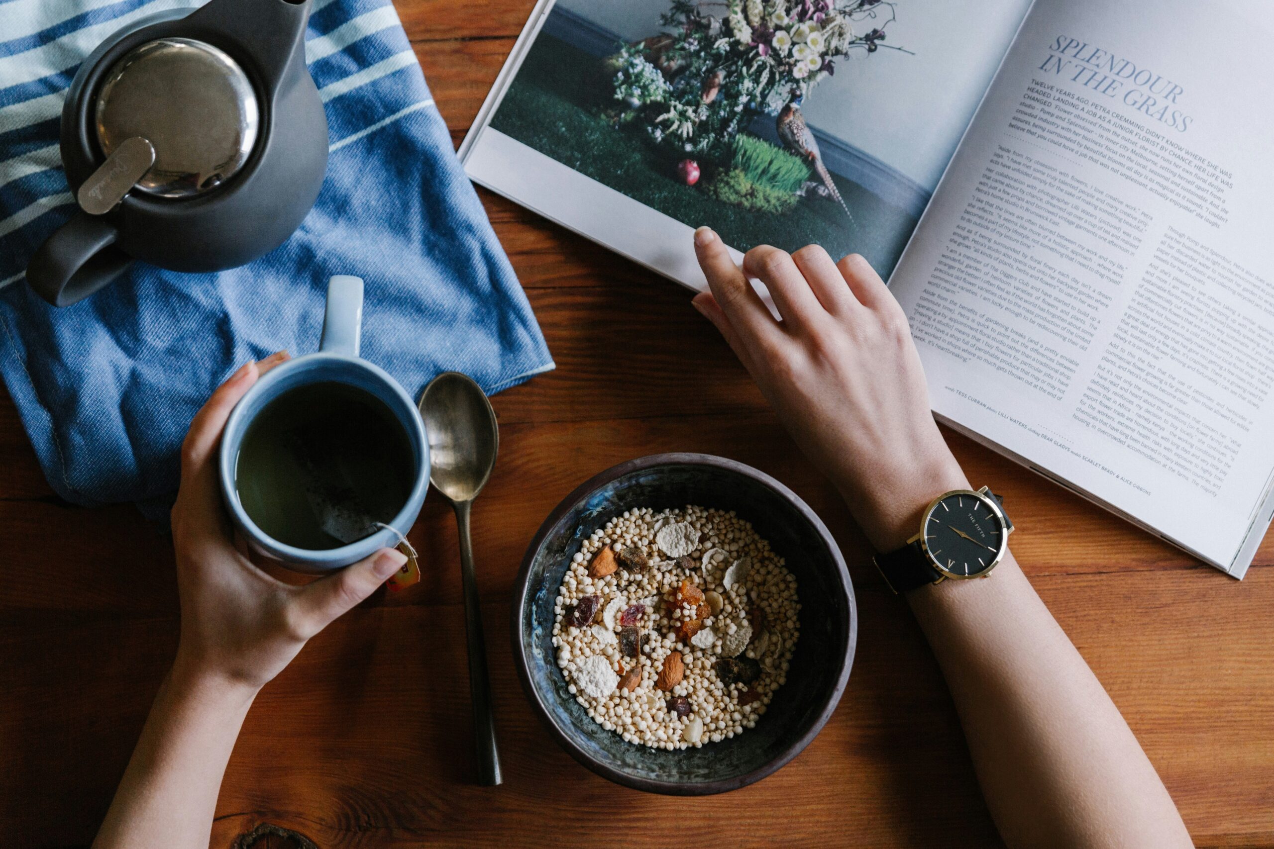 A person eating a healthy breakfast of cereal with nuts and dried fruit while reading a magazine, with a cup of tea and teapot nearby on a wooden table.